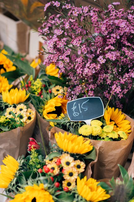 A vibrant display of fresh floral arrangements featuring bright yellow sunflowers with large, open petals and green foliage, wrapped in brown craft paper. Interspersed among the sunflowers are smaller