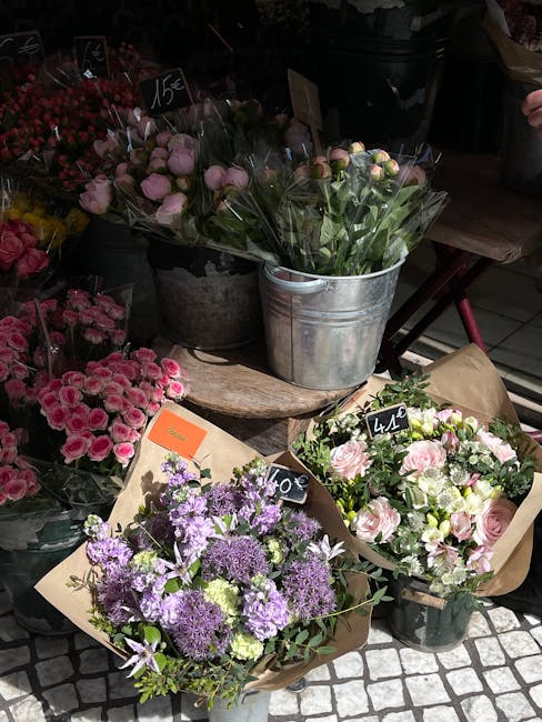 A display of fresh flower bouquets at a flower shop, arranged in various containers including a galvanized metal bucket and wrapped in brown paper. The bouquets feature a variety of flowers in soft pa