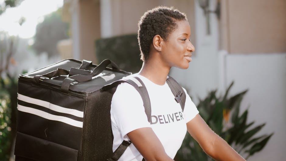A young woman with short curly hair and medium skin tone, dressed in a white T-shirt with the word 'DELIVERY' printed on it, is outdoors during daylight. She is carrying a large, black insulated deliv