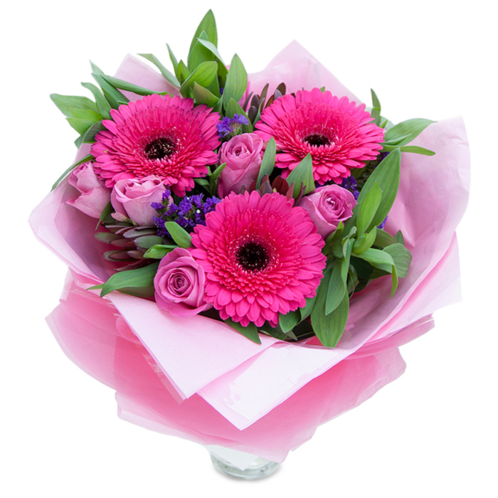 This bouquet reads like a small, joyful statement in pinks and purples, set simply against a white background so every colour and texture can be appreciated. Three confident Gerbera daisies in a saturated fuchsia dominate the eye - their broad, evenly spaced petals radiate outward toward centres that are nearly black, providing a striking, modern contrast. Tucked among these are roses in a muted lavender-pink or dusty rose hue, their petals still forming tight, velvety spirals that evoke both romance and freshness. Little clouds of purple statice or limonium add a papery, textural punctuation, drifting between the larger blooms and softening transitions. The foliage is generous and glossy, likely salal and eucalyptus, their cool green tones and slightly waxy textures supporting the bouquet's silhouette and adding a hint of crisp, leafy scent. The wrapping is deliberately elegant: an inner layer of soft matte pale pink tissue creates a gentle base while an outer sheet of translucent fuchsia-pink cellophane with a faint crinkle produces a subtle sheen, folded with practiced hands to frame the flowers. The clear glass vase rim visible at the bottom signals that the arrangement is ready to be placed on display. In natural daylight the composition feels uplifting yet intimate - the roses offer a soft perfume, the Gerberas contribute a faint green sweetness, and the entire arrangement conveys affection and celebration. It's the sort of bouquet a local Clerkenwell florist might prepare for deliveries around St John Street or visitors arriving from nearby Smithfield Market - a thoughtful, handcrafted gift for birthdays, anniversaries, or a spontaneous expression of care.